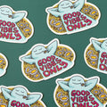 A blue, cartoon character with arms outstretched is lying on its back in a field of daisies. The text "GOOD VIBES ONLY" appears in bold, colorful letters below the character.