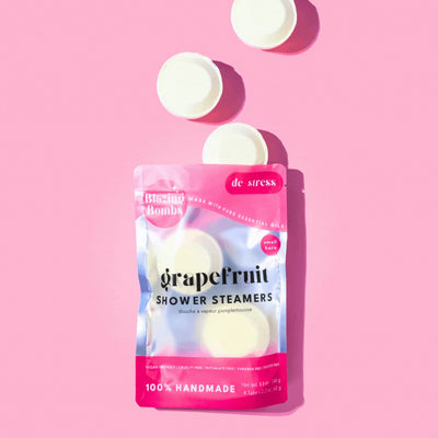 A vibrant, high-angle flat lay shot on a pink background features a shiny, pink and silver pouch of Blazing Bombs Grapefruit Shower Steamers. The pouch is labeled 