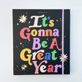 A black agenda with the phrase "It's Gonna Be A Great Year" written in colorful letters. The letters are outlined in gold and there are gold stars scattered around the text. The agenda has a purple elastic band.