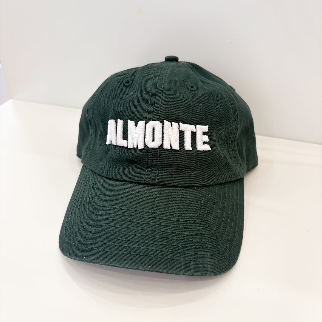 A dark green cotton baseball cap with 'ALMONTE' embroidered in white block letters across the front, displayed on a white surface against a white background.