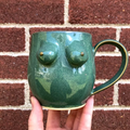 Green ceramic mug with breast-like design held against a brick wall.