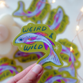 Hand holding a colorful sticker with a fish design and 'WEIRD & WILD' text, surrounded by more stickers.