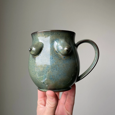 Blue Boob Mug, a big coffee cup with large handle with 3 dimensional breasts on the front, in a blue green colour.