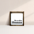 A framed square wooden sign with the text "Be a nice Human" in bold, black letters, centered on a white background. The frame is a dark brown wood with a rustic finish.