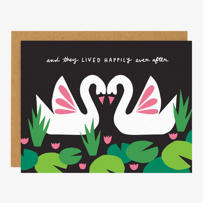 A black greeting card with a brown envelope. The card features two white swans with pink wings facing each other in a heart shape. They are surrounded by green lily pads and pink flowers. The text 