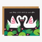 A black greeting card with a brown envelope. The card features two white swans with pink wings facing each other in a heart shape. They are surrounded by green lily pads and pink flowers. The text 