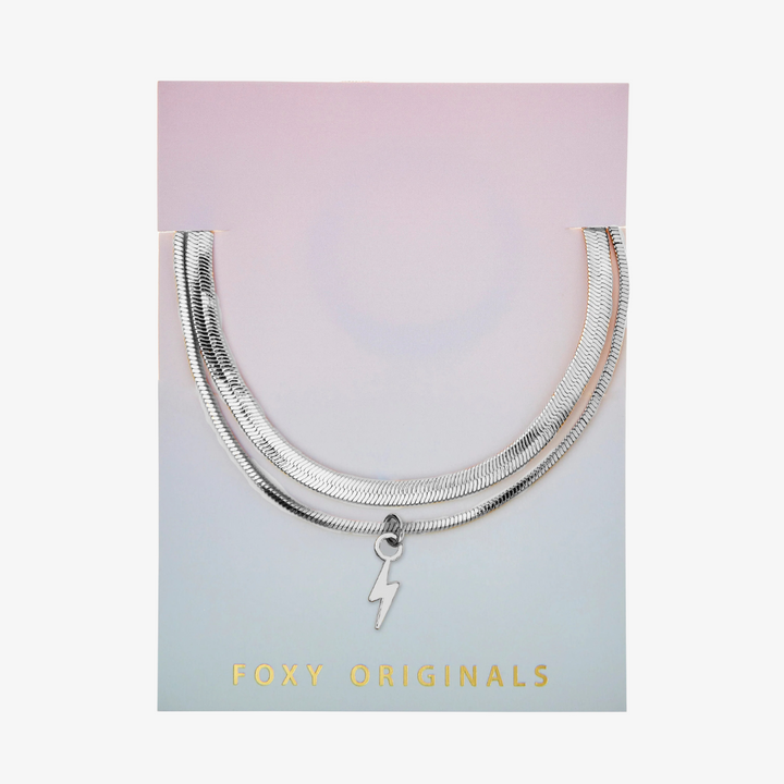 A set of two silver bracelets, one layered over the other, with a lightning bolt charm hanging from the bottom. The bracelets are displayed on a pink card with the brand name 