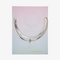 A set of two silver bracelets, one layered over the other, with a lightning bolt charm hanging from the bottom. The bracelets are displayed on a pink card with the brand name 