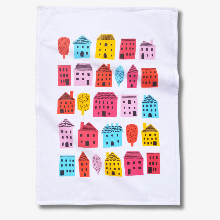 A white tea towel with a pattern of colorful houses and trees. The houses are in various sizes and colors, including pink, red, orange, yellow, blue, and green. The trees are green and have simple leaf shapes.