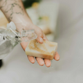A rectangular bar of shaving soap, wrapped in a paper band with the text "Zero Waste MVMT Shave Bar Blossom". The soap is a light brown color with a creamy texture, and the seed paper band is white with a leaf illustration.