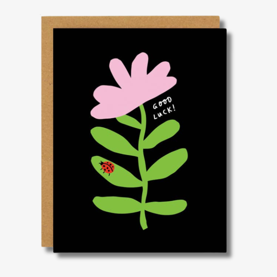 Good Luck Ladybug Encouragement Card. A simple, playful design featuring a large pink flower with green leaves on a black background. A red ladybug is perched on one of the leaves, and the text 'Good Luck!' is written in white next to the flower. A kraft paper envelope is partially visible behind the card, adding a rustic touch.