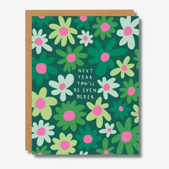 Next Year Birthday Card: A humorous birthday card featuring a dark green background with stylized white, light green, and pink flowers. In the center, white text reads ‘NEXT YEAR YOU’LL BE EVEN OLDER.’