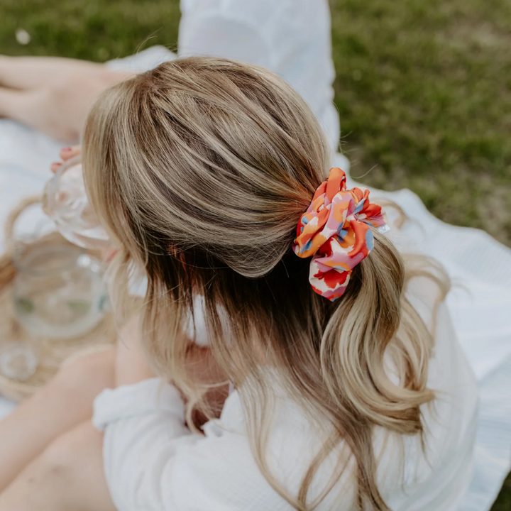 Flower Child Petite Scunchie, a fluffy hair tie in a rainbow of tiedye colours in a blonde woman's hair.