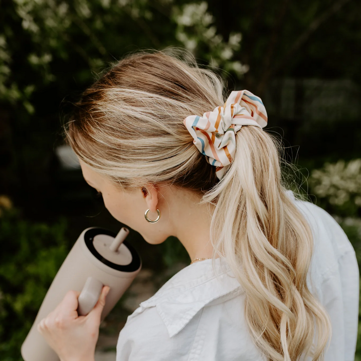 Sundeck Stripes Petite Scunchie, a fluffy hair tie in a creamy white with a rainbow of stripes in a blonde woman's hair.