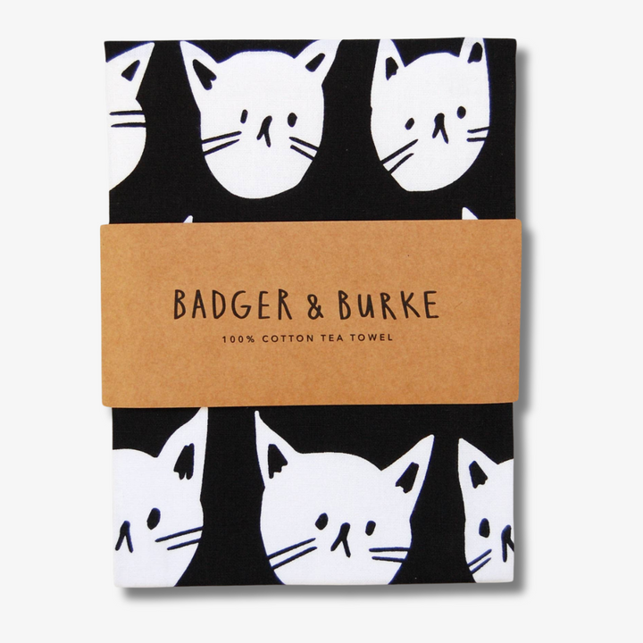 A black tea towel with a pattern of white cat faces. The cat faces are arranged in a grid-like pattern and vary in size and shape.