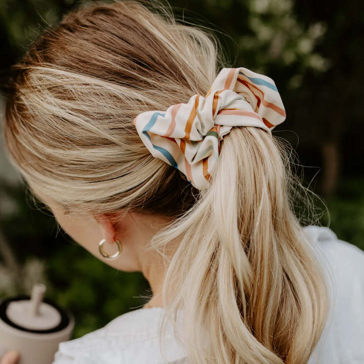 Sundeck Stripes Petite Scunchie, a fluffy hair tie in a creamy white with a rainbow of stripes in a blonde woman's hair.