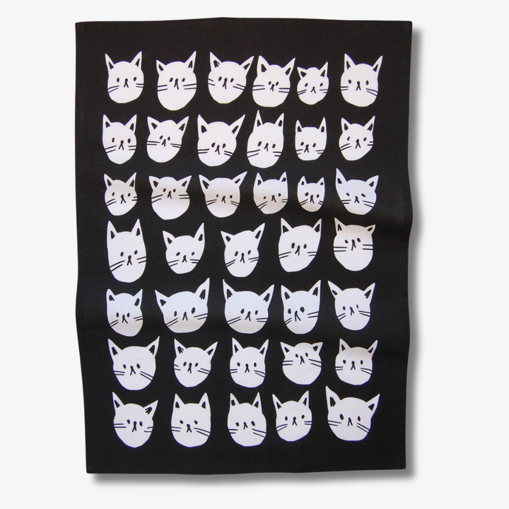 A black tea towel with a pattern of white cat faces. The cat faces are arranged in a grid-like pattern and vary in size and shape.