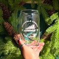 Clear glass can-shaped tumbler with engraved design held in front of a Christmas tree.