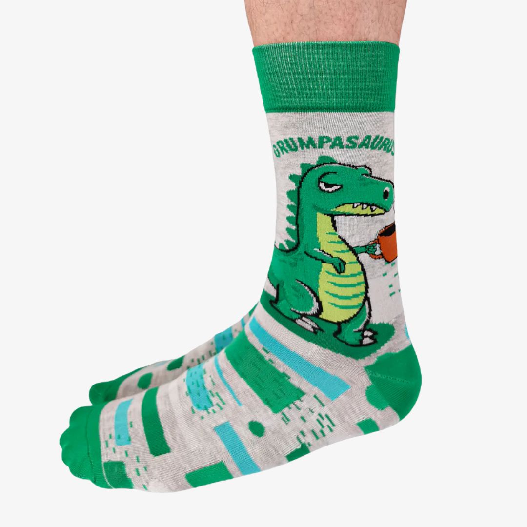 A pair of grey crew socks with horizontal stripes in shades of green and blue. One sock features a cartoon illustration of a grumpy green dinosaur holding a brown coffee mug and the text "GRUMPASAURUS" in green letters above it. The cuff, heel, and toe of the sock are a darker shade of green. The brand "Uptown Sox" is subtly visible on the sole.