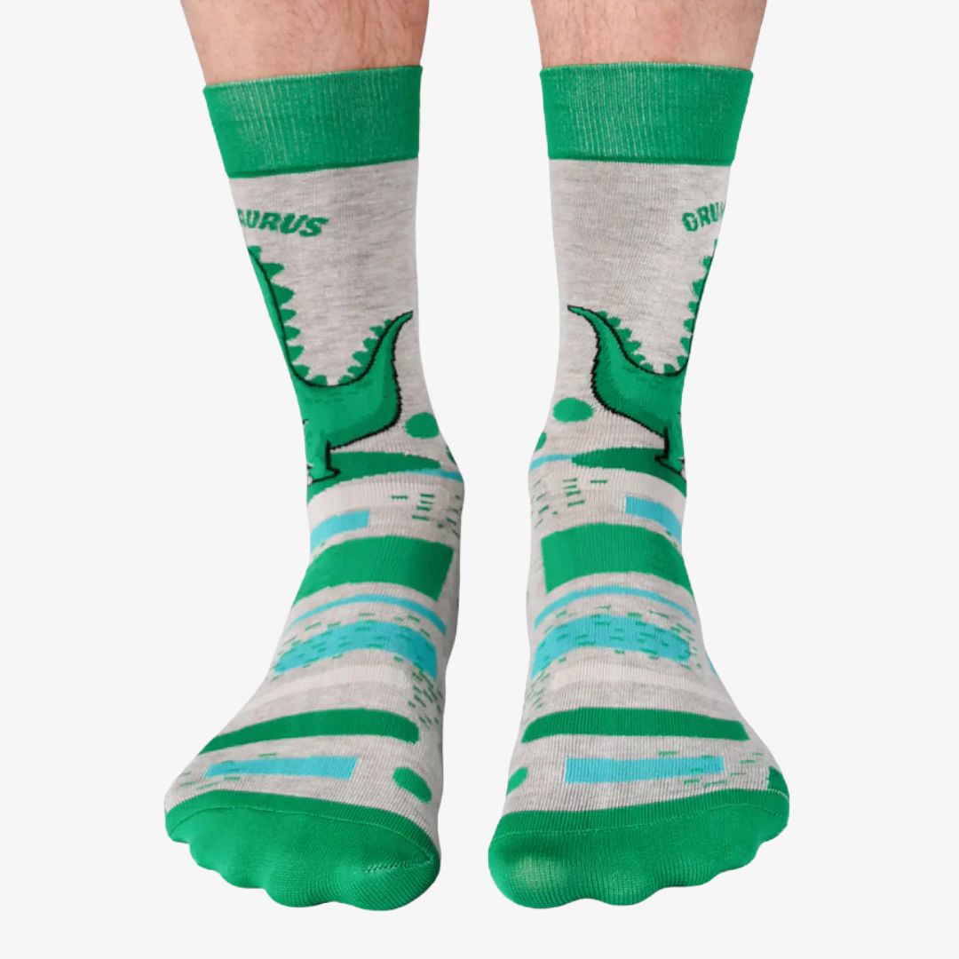A pair of grey crew socks with horizontal stripes in shades of green and blue. One sock features a cartoon illustration of a grumpy green dinosaur holding a brown coffee mug and the text "GRUMPASAURUS" in green letters above it. The cuff, heel, and toe of the sock are a darker shade of green. The brand "Uptown Sox" is subtly visible on the sole.