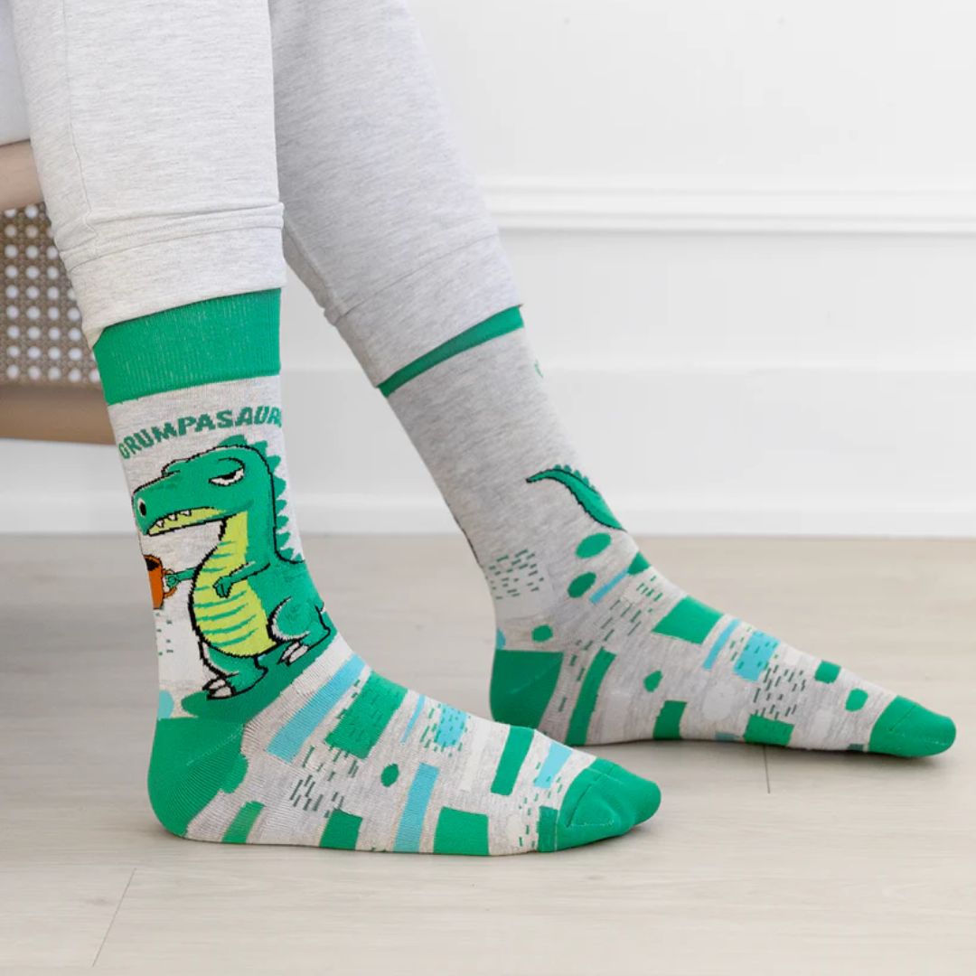 A pair of grey crew socks with horizontal stripes in shades of green and blue. One sock features a cartoon illustration of a grumpy green dinosaur holding a brown coffee mug and the text "GRUMPASAURUS" in green letters above it. The cuff, heel, and toe of the sock are a darker shade of green. The brand "Uptown Sox" is subtly visible on the sole.