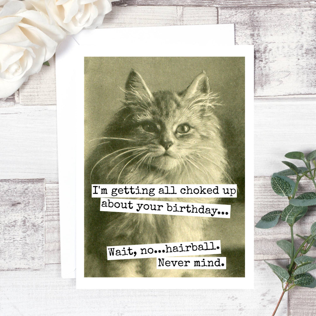 A birthday card featuring a vintage black and white photo of a fluffy cat looking directly forward. Text on the card reads: "I'm getting all choked up about your birthday... Wait, no...hairball. Never mind."