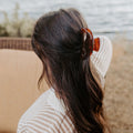 Person with long hair wearing a brown hair clip, sitting by a body of water.