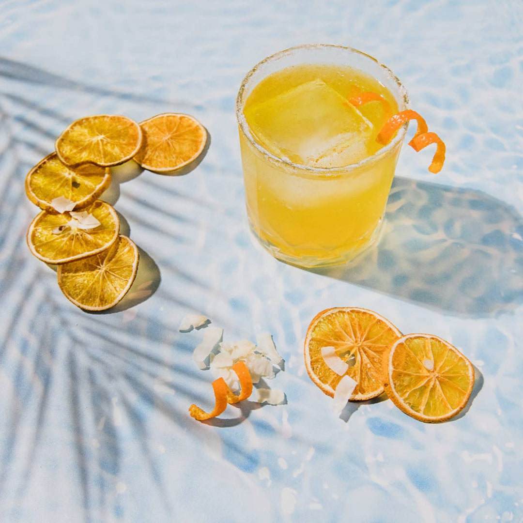 Glass of yellow cocktail with ice and orange slices on a light blue surface with palm leaf