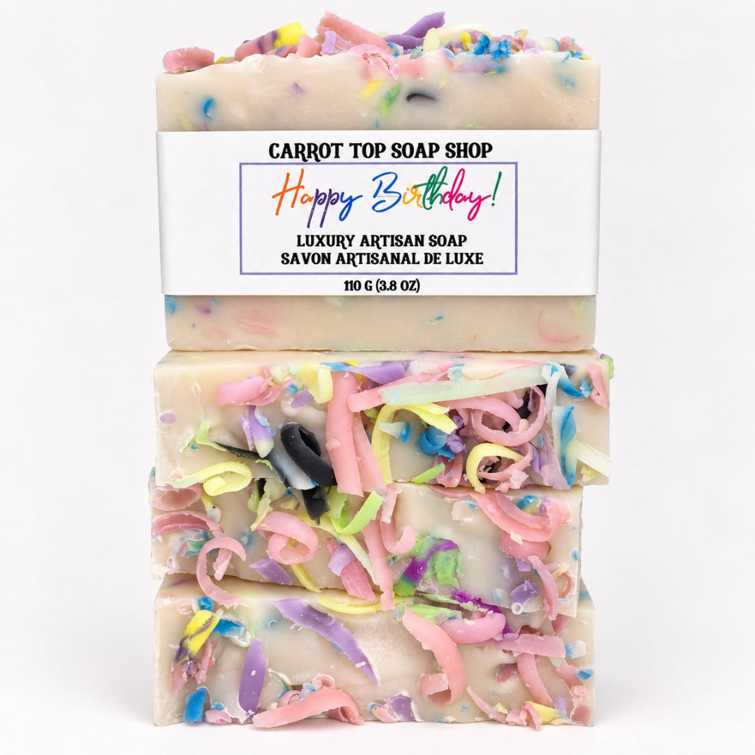 Three stacked bars of artisanal soap with a white paper label reading 'CARROT TOP SOAP SHOP Happy Birthday! Luxury Artisan Soap'. The soap is creamy off-white with colorful curly shavings and multicolored flecks throughout, photographed on a clean white background.