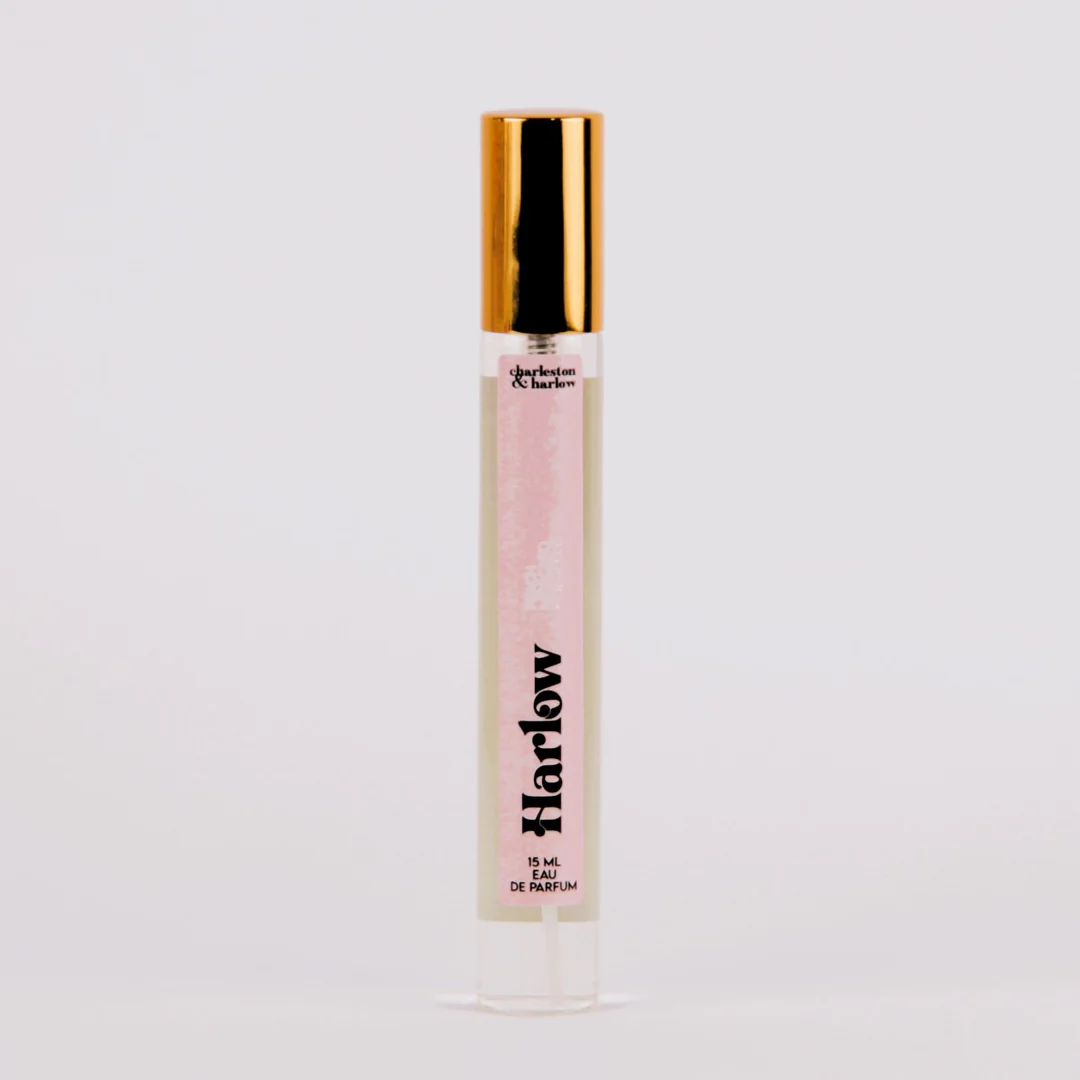 A bottle of Charleston & Harlow perfume named "Harlow." The bottle is clear glass with a gold cap. The label is pink with the brand name and perfume name in a bold, playful font. The fragrance notes are listed as "Peach, Moscato, Pineapple."
