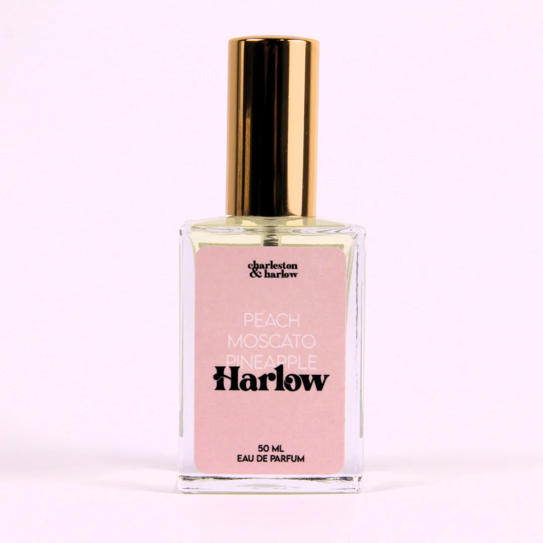 A bottle of Charleston & Harlow perfume named "Harlow." The bottle is clear glass with a gold cap. The label is pink with the brand name and perfume name in a bold, playful font. The fragrance notes are listed as "Peach, Moscato, Pineapple."