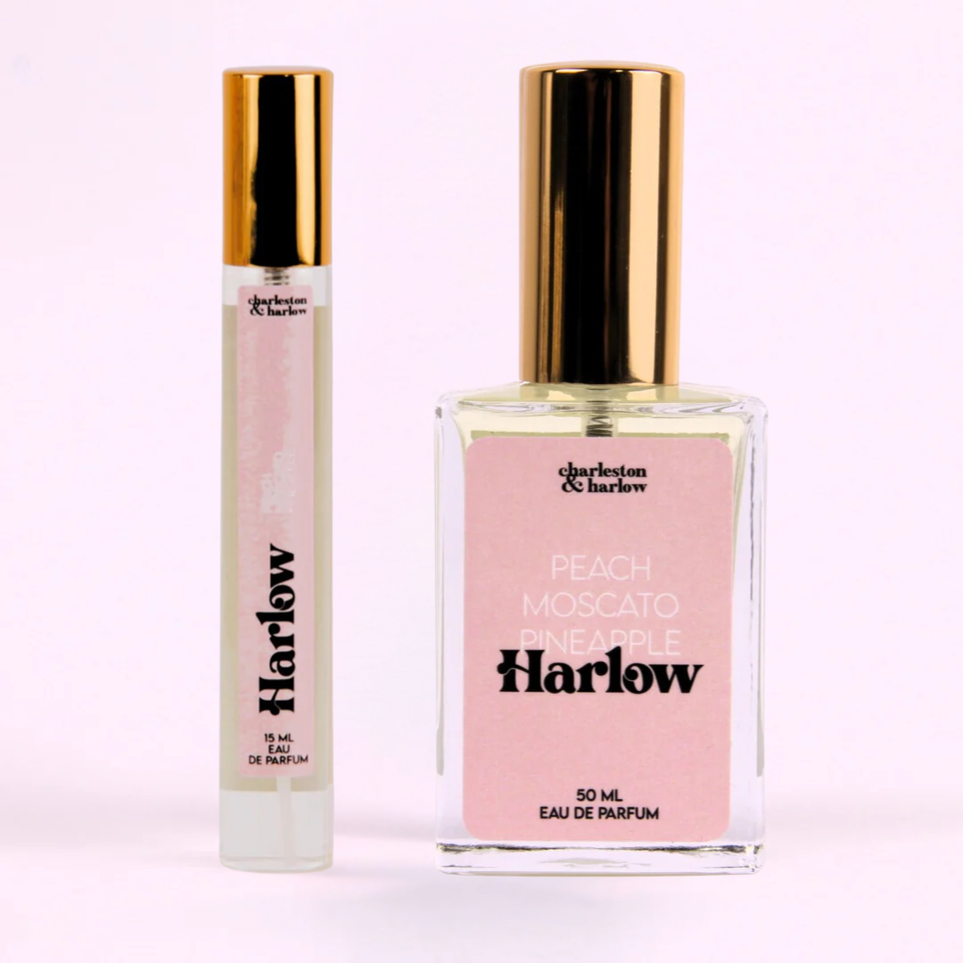 A bottle of Charleston & Harlow perfume named "Harlow." The bottle is clear glass with a gold cap. The label is pink with the brand name and perfume name in a bold, playful font. The fragrance notes are listed as "Peach, Moscato, Pineapple."