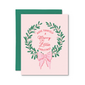 A square Christmas greeting card with a pale pink background, featuring a green leaf wreath and pink script text that reads 'HAVE YOURSELF A Merry Little CHRISTMAS,' with a pink bow illustration below. A dark green envelope is placed behind the card on a white background.