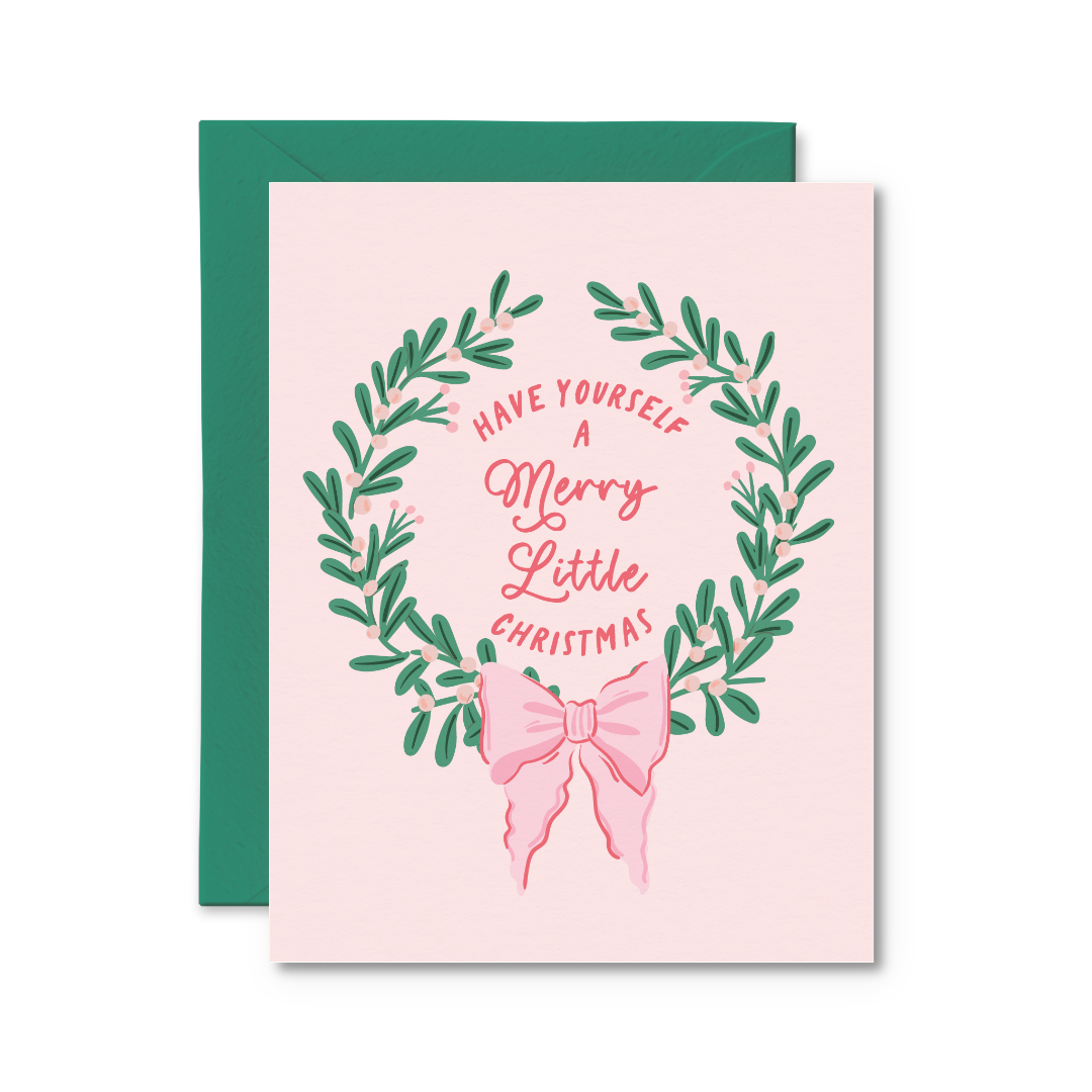 A square Christmas greeting card with a pale pink background, featuring a green leaf wreath and pink script text that reads 'HAVE YOURSELF A Merry Little CHRISTMAS,' with a pink bow illustration below. A dark green envelope is placed behind the card on a white background.