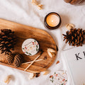 Wooden tray with pinecones, a candle, and a small round container on a white surface.