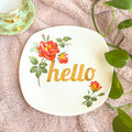Round white plate with floral designs and 'hello' text on a textured surface with leaves.