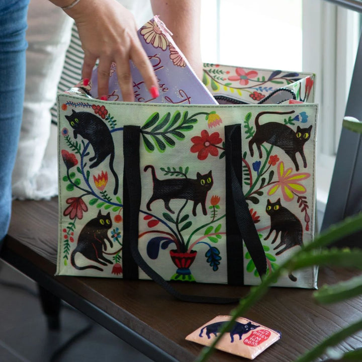 A rectangular shoulder tote bag with black handles, featuring a whimsical pattern of black cats, colourful flowers, and foliage against a white background.