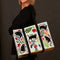 A rectangular shoulder tote bag with black handles, featuring a whimsical pattern of black cats, colourful flowers, and foliage against a white background.