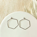 A pair of thin silver-tone hexagonal hoop earrings resting on a white ceramic plate, with a light beige fabric background.
