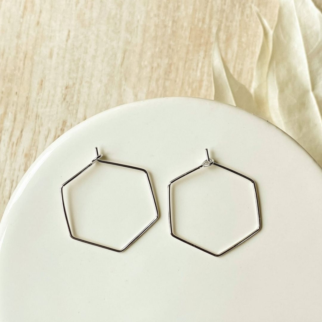 A pair of thin silver-tone hexagonal hoop earrings resting on a white ceramic plate, with a light beige fabric background.