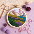Embroidery hoop with a colorful landscape design on a textured surface with fabric and yarn.