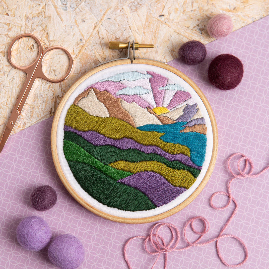 Embroidery hoop with a colorful landscape design on a textured surface with fabric and yarn.