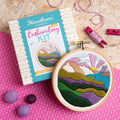 Embroidery kit with completed mountain landscape design on a pink fabric background