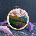Embroidery hoop with a colorful mountain landscape design on a dark blue background.