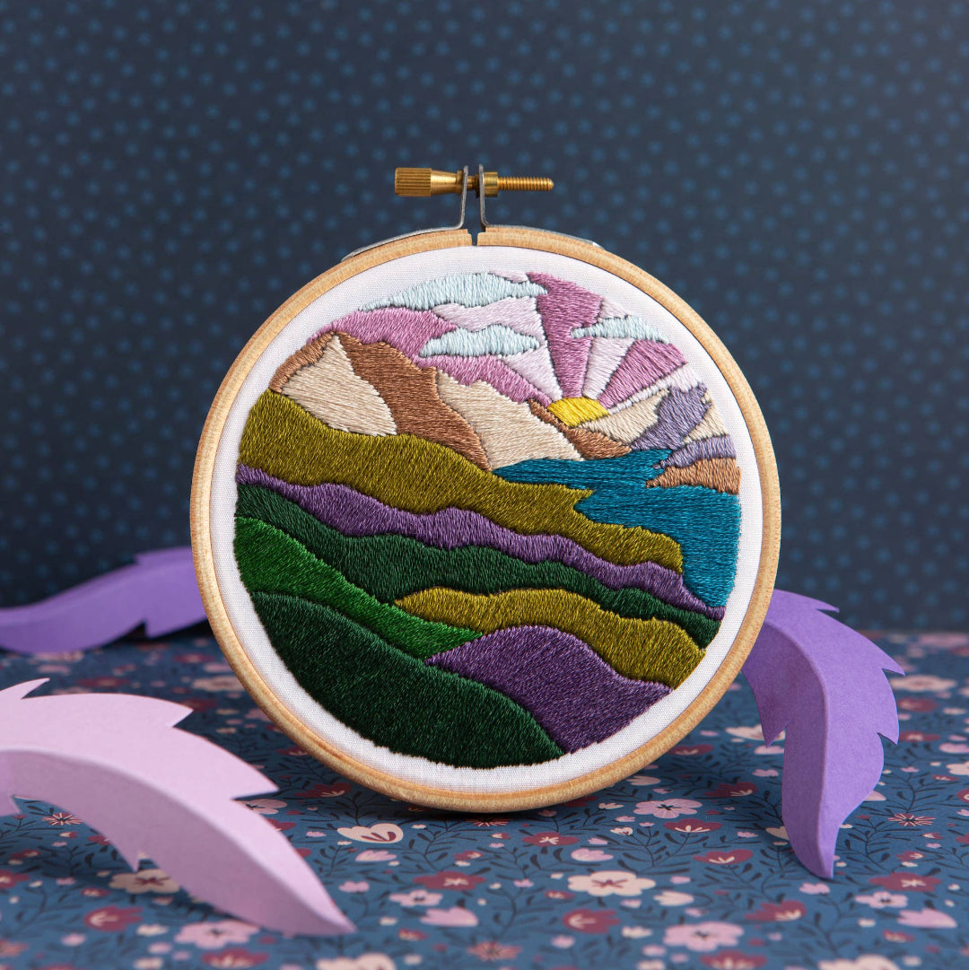 Embroidery hoop with a colorful mountain landscape design on a dark blue background.