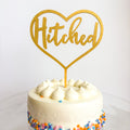 White cake with colorful sprinkles and a gold 'Hitched' heart-shaped topper on a white background