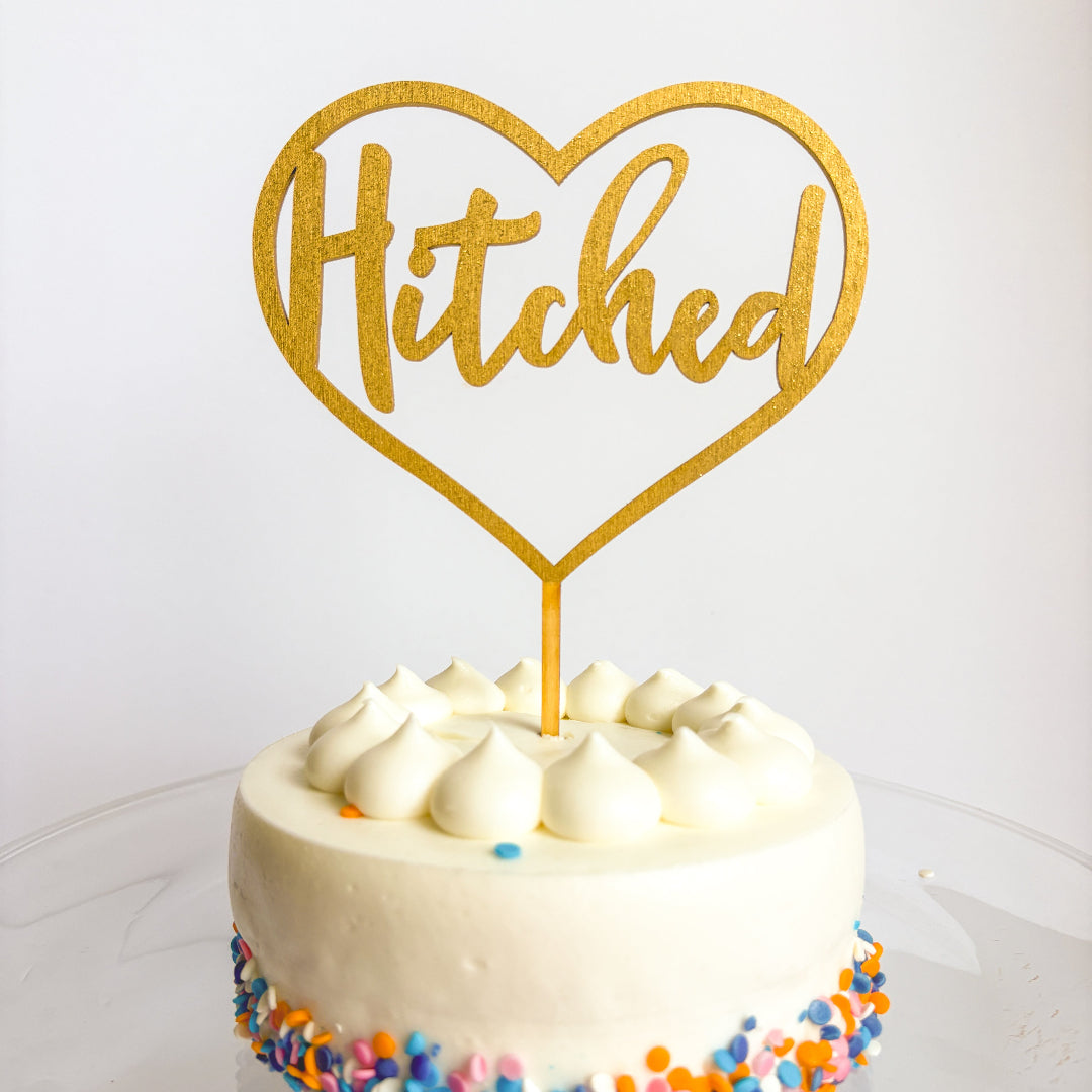 White cake with colorful sprinkles and a gold 'Hitched' heart-shaped topper on a white background