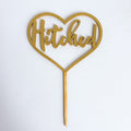Gold heart-shaped cake topper with 'Hitched' text on a white background