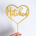 Gold heart-shaped cake topper with 'Hitched' text on a white background