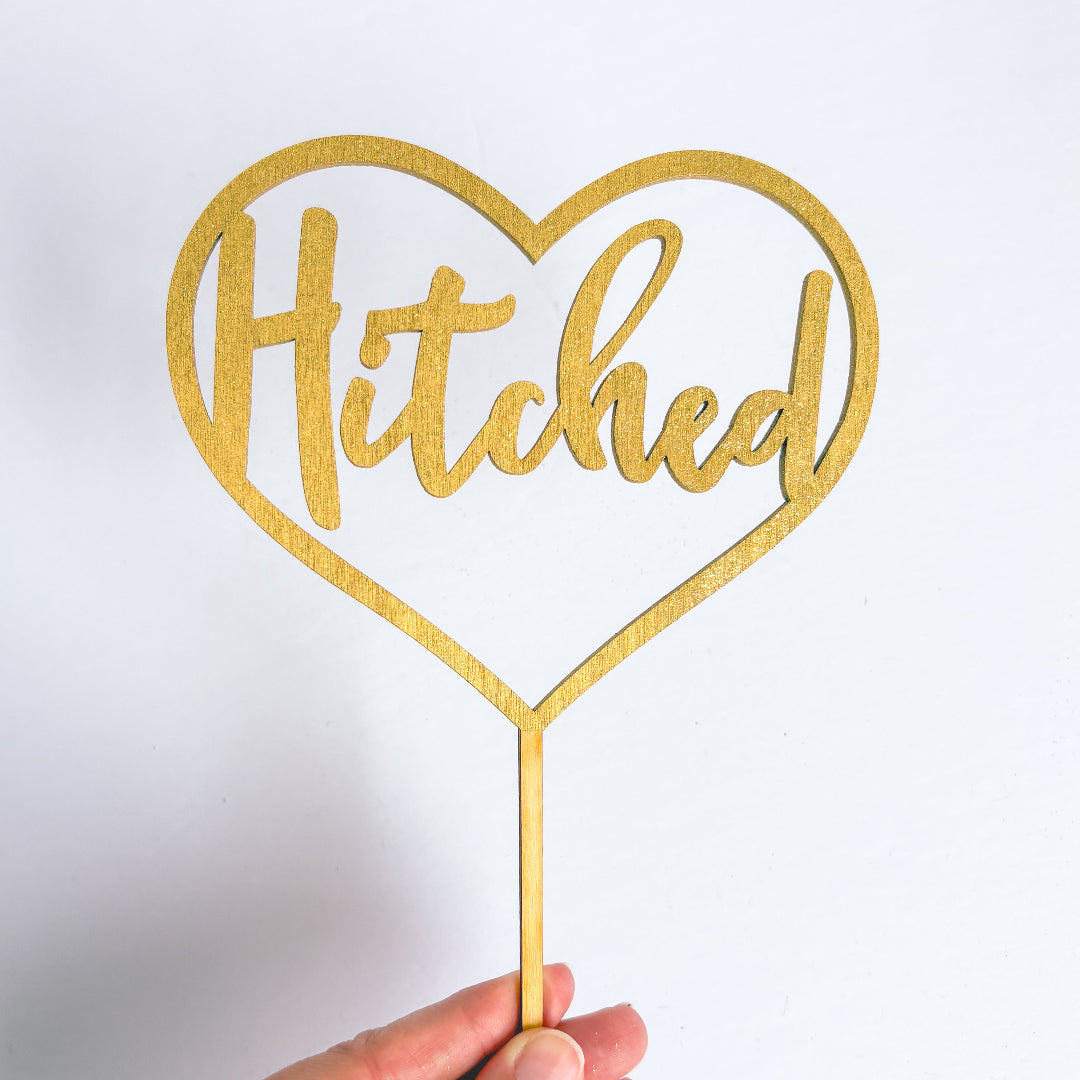 Gold heart-shaped cake topper with 'Hitched' text on a white background